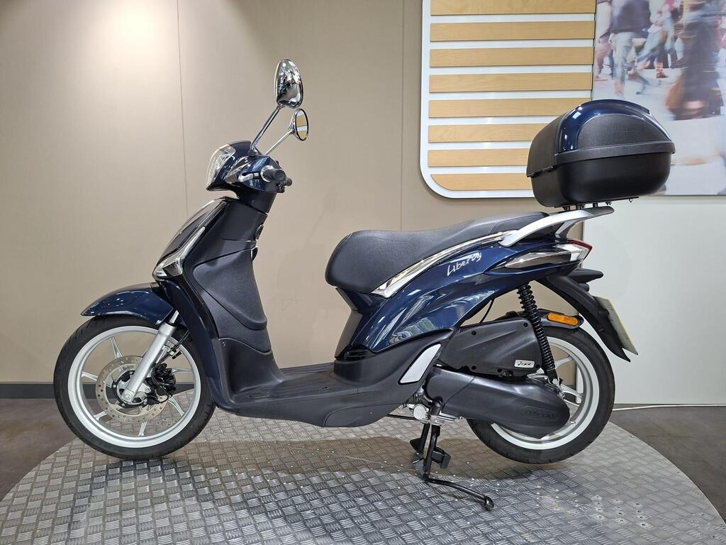 Piaggio Liberty 50 (2019) 15 A blue scooter with a sleek design and a rear storage compartment, displayed on a textured surface against a light-colored wall.