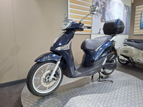 A blue scooter is displayed on a circular metal platform in a showroom, showcasing its sleek design, front fender, and rear storage box.