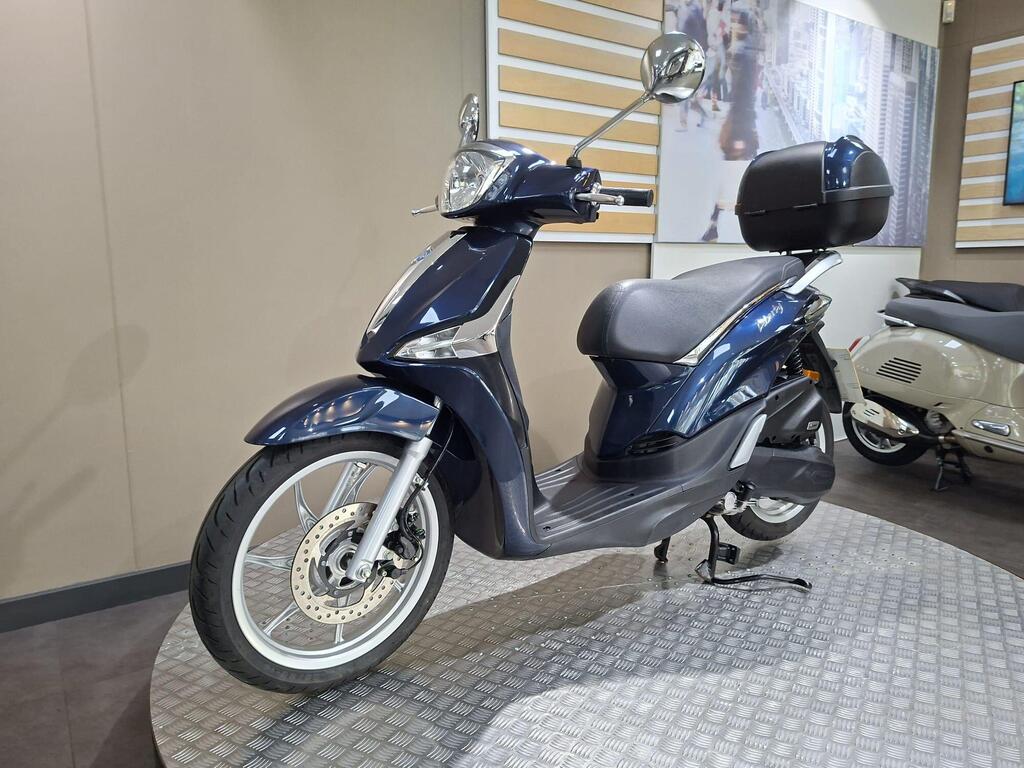 Piaggio Liberty 50 (2019) 16 A blue scooter is displayed on a circular metal platform in a showroom, showcasing its sleek design, front fender, and rear storage box.