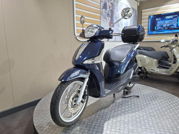 A blue scooter displayed on a circular platform in a showroom, with a digital screen showing information in the background.