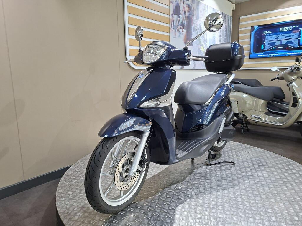 Piaggio Liberty 50 (2019) 17 A blue scooter displayed on a circular platform in a showroom, with a digital screen showing information in the background.