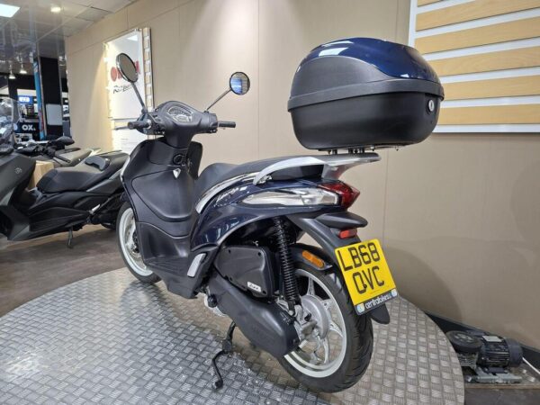 A blue and silver scooter is parked on a metal platform inside a showroom, featuring a top case. The back license plate reads "LB68 OVC." Other scooters are visible in the background.