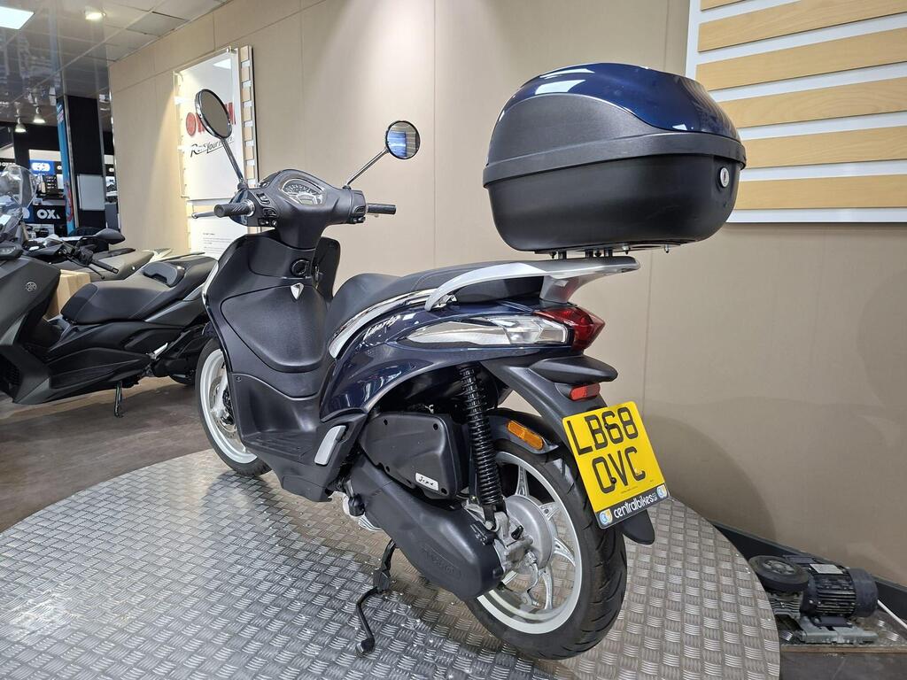 Piaggio Liberty 50 (2019) 18 A blue and silver scooter is parked on a metal platform inside a showroom, featuring a top case. The back license plate reads "LB68 OVC." Other scooters are visible in the background.