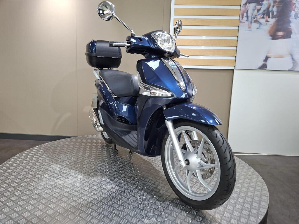 Piaggio Liberty 50 (2019) 19 A blue scooter displayed on a round platform with a textured surface, featuring a rounded front, a single rearview mirror, and a black storage box attached to the back. The background includes a light-colored wall and a large image of people walking.