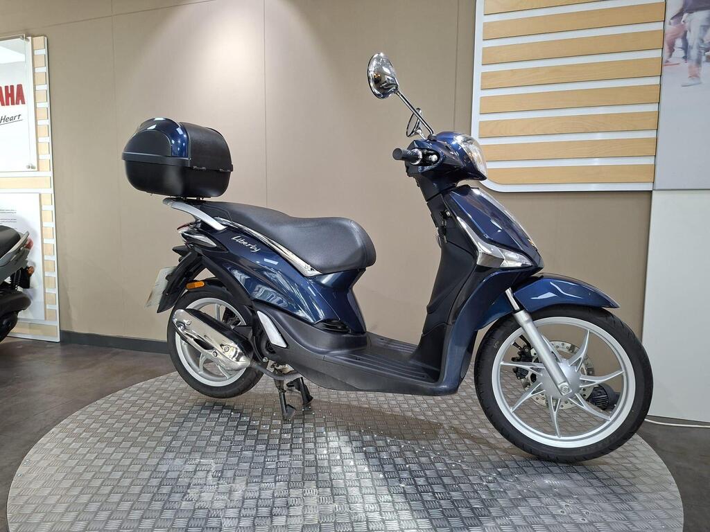 Piaggio Liberty 50 (2019) 2 A blue scooter with a black top case is displayed on a textured round platform in a showroom. The background features a neutral-colored wall and Yamaha branding.