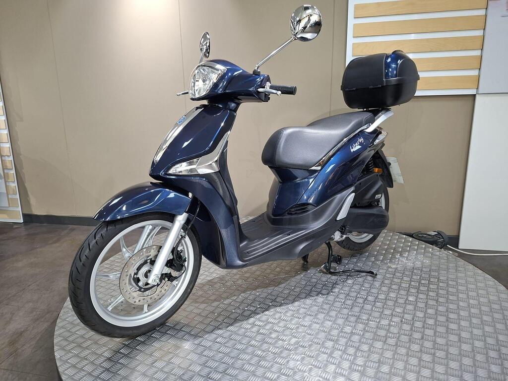 Piaggio Liberty 50 (2019) 20 A navy blue scooter with a storage compartment on the back, displayed on a circular platform with a textured surface, set against a neutral-colored wall.