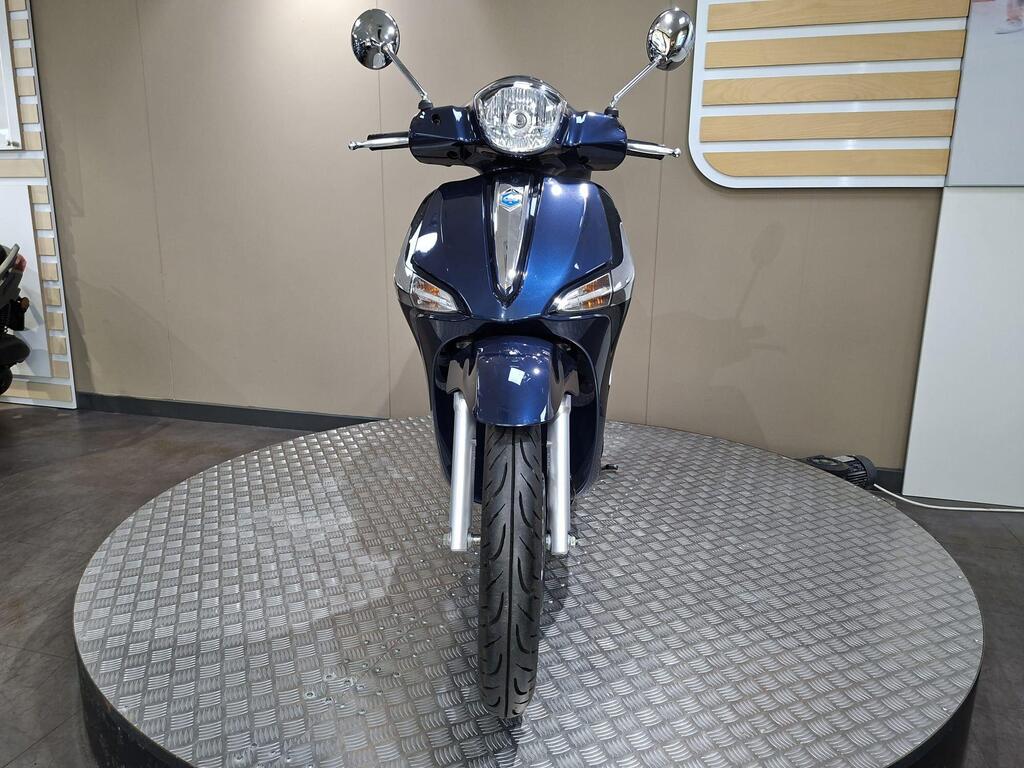 Piaggio Liberty 50 (2019) 22 A blue scooter is displayed on a circular platform, viewed from the front, showcasing its sleek design, chrome mirrors, and detailed rim pattern. The background features a neutral-colored wall and visible reflections.