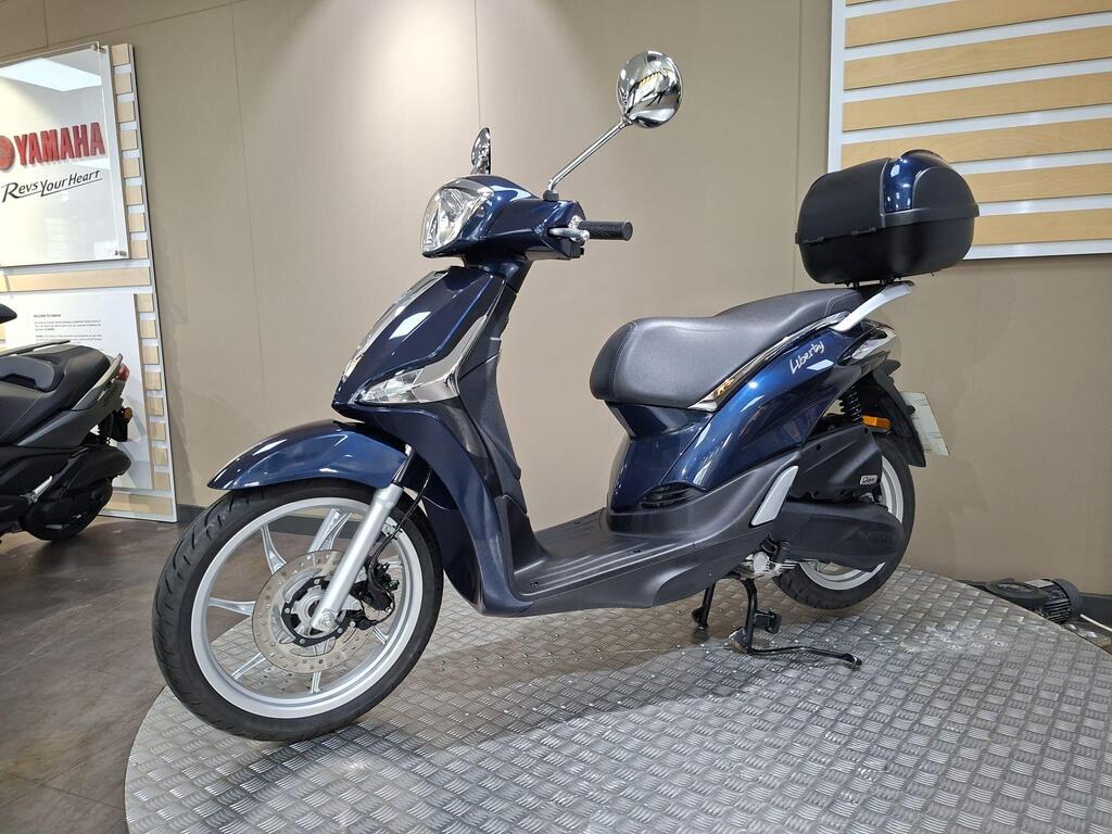 Piaggio Liberty 50 (2019) 23 A blue Yamaha scooter displayed on a circular platform, featuring a stylish design, a comfortable seat, a rear storage box, and shiny alloy wheels, set against a neutral background with Yamaha branding visible.