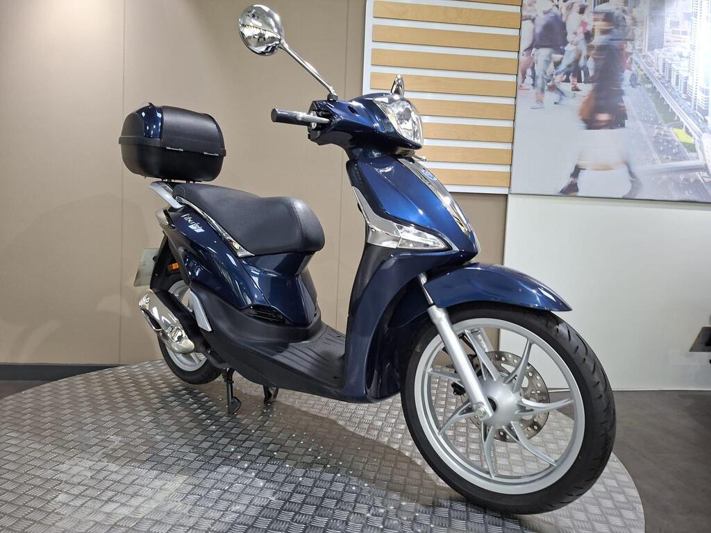 Piaggio Liberty 50 (2019) 24 A blue scooter is displayed on a textured platform, featuring a comfortable seat and a black storage compartment on the back, with a backdrop of a blurred urban setting.
