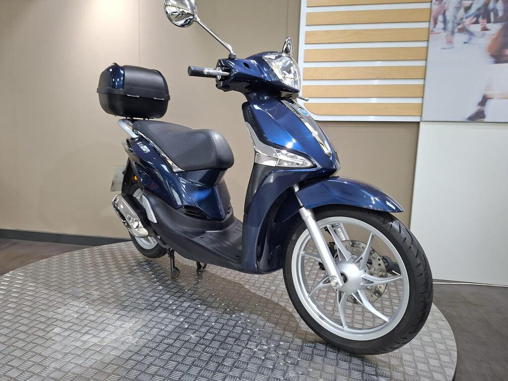 Piaggio Liberty 50 (2019) 25 A blue scooter with a black storage box on the rear, positioned on a textured surface, against a neutral background.