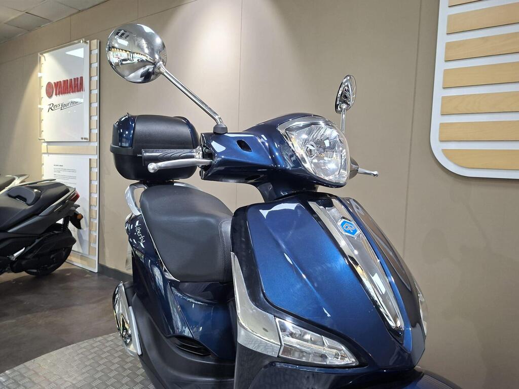 Piaggio Liberty 50 (2019) 26 A close-up view of a blue scooter with a shiny front headlight and rearview mirrors, displayed in a showroom with a Yamaha logo visible in the background.