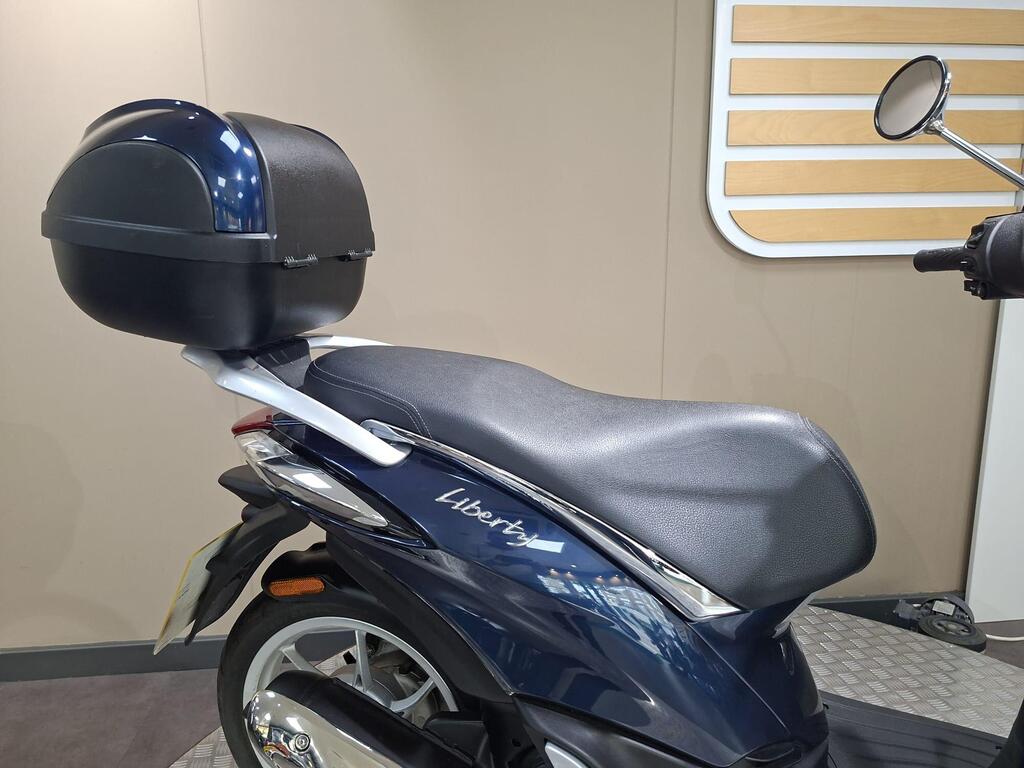 Piaggio Liberty 50 (2019) 27 A close-up side view of a blue scooter featuring a comfortable black seat and a rear storage box, set against a neutral background with horizontal wooden slats.