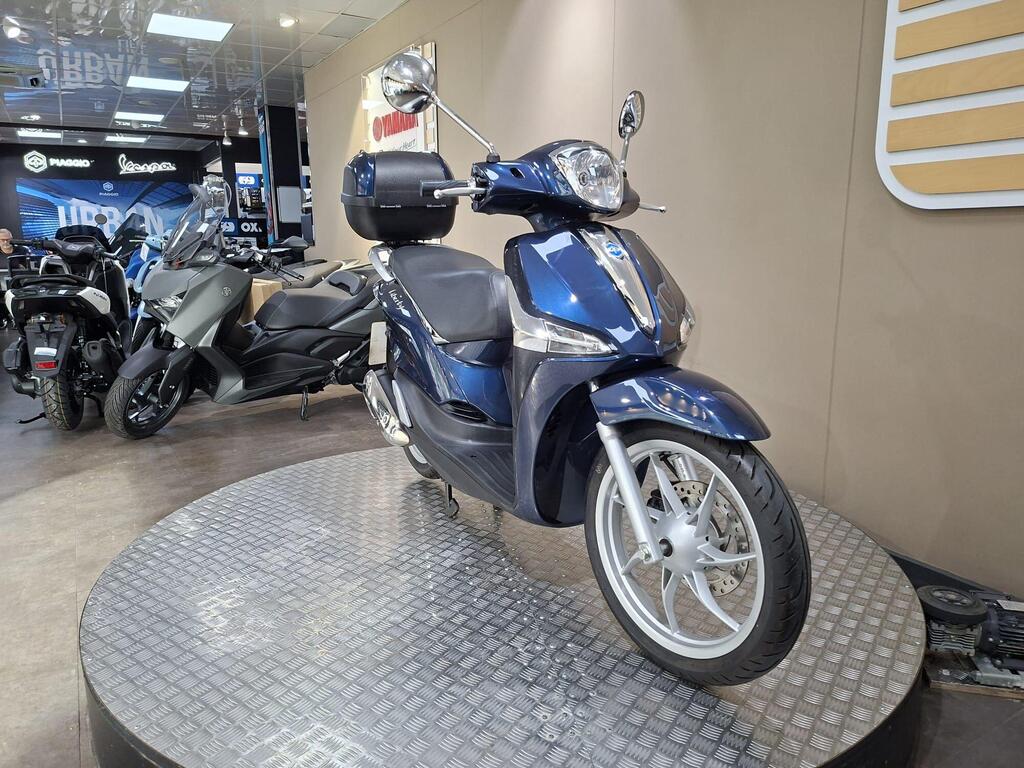 Piaggio Liberty 50 (2019) 3 A blue scooter displayed on a circular platform in a motorcycle showroom, with other scooters and motorcycles visible in the background.