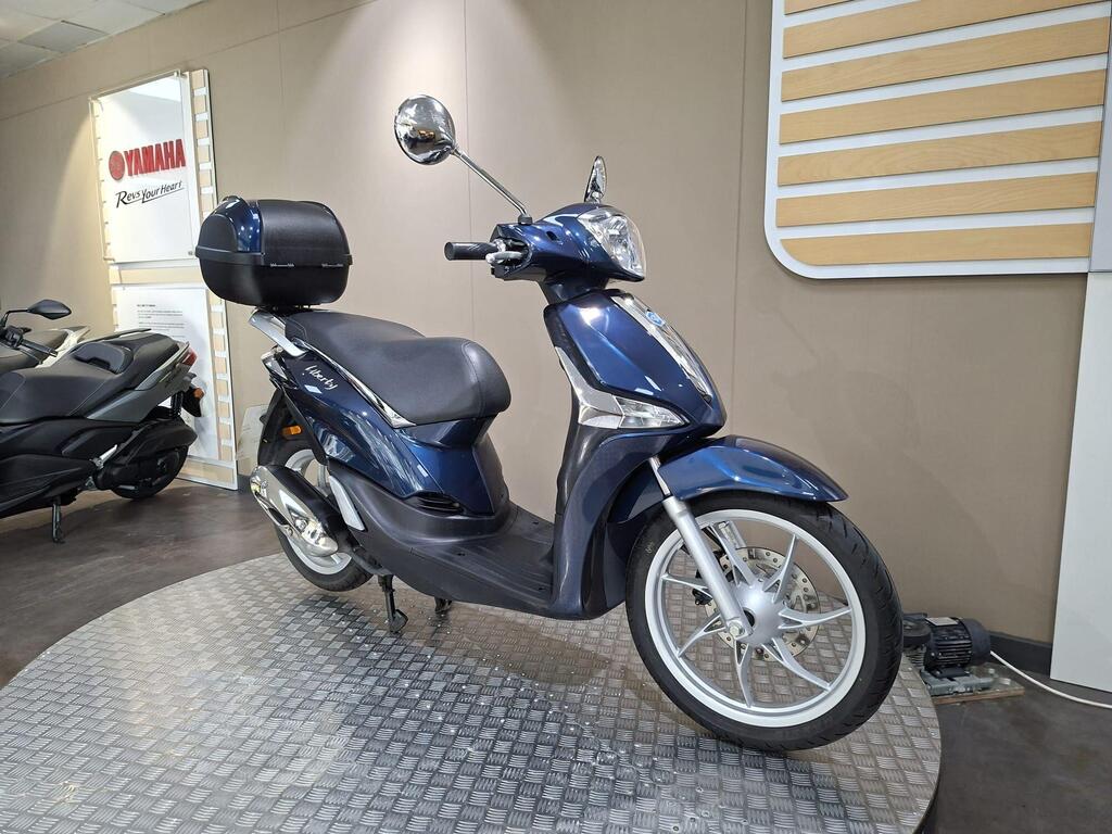 Piaggio Liberty 50 (2019) 4 A blue scooter is displayed on a circular platform in a showroom, featuring a comfortable seat, a rear storage compartment, and chrome accents, with a minimalist background.