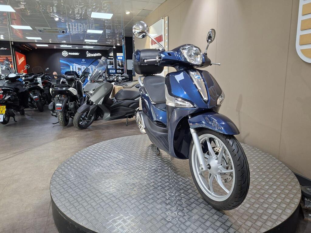 Piaggio Liberty 50 (2019) 5 A blue scooter displayed on a round platform in a motorcycle showroom, with various other motorcycles visible in the background.