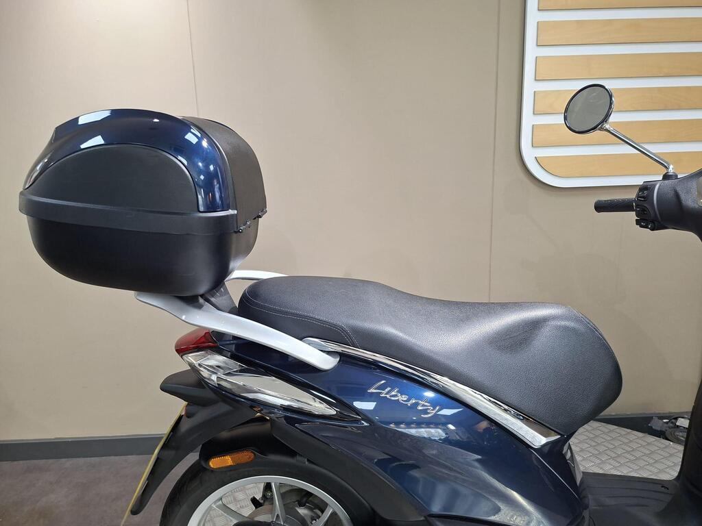 Piaggio Liberty 50 (2019) 6 A close-up view of a navy blue scooter with a black top case, featuring a textured seat and chrome detailing on the side, set against a neutral background.