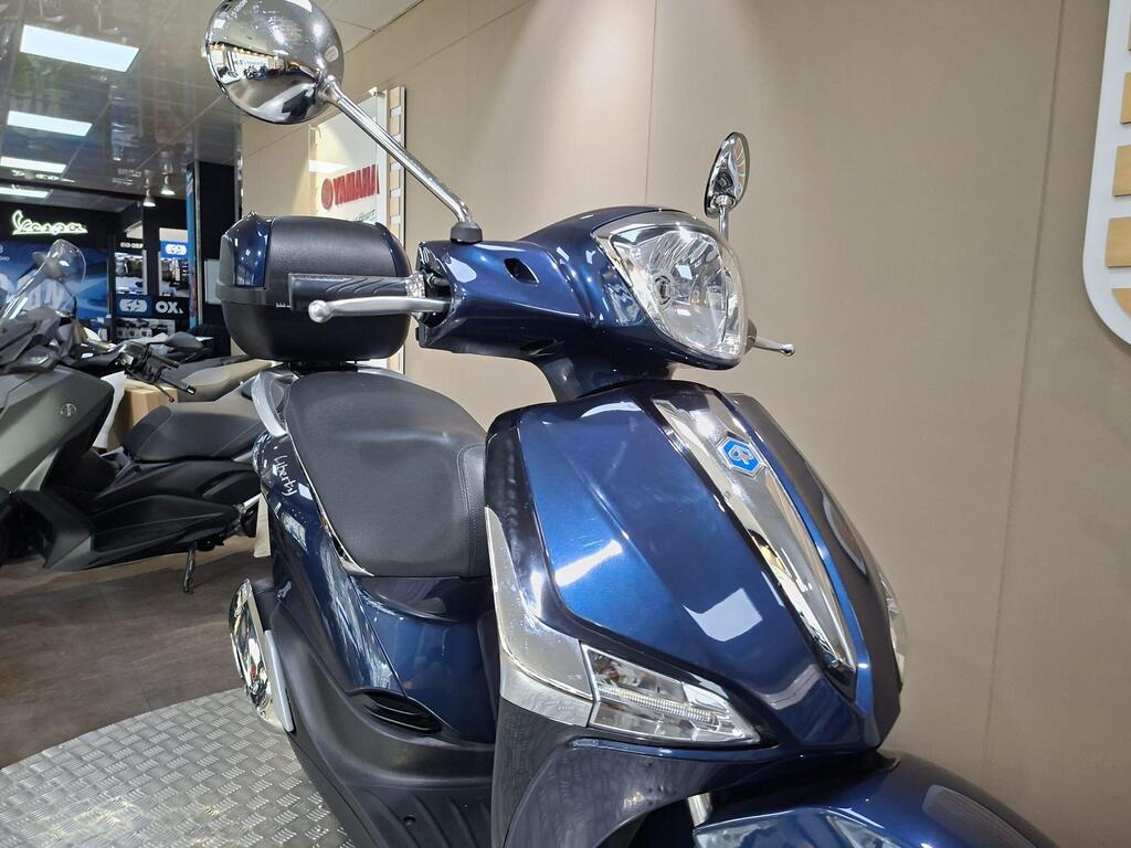 Piaggio Liberty 50 (2019) 7 A close-up view of a blue scooter featuring a sleek design, prominent headlight, and rearview mirror, displayed in a modern showroom.