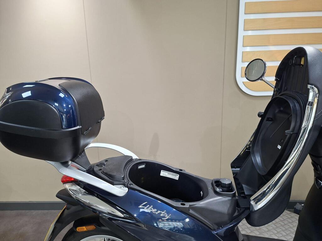 Piaggio Liberty 50 (2019) 8 Close-up of a blue scooter with a black storage compartment and rear trunk, showcasing the open seat and storage area, set against a neutral backdrop.