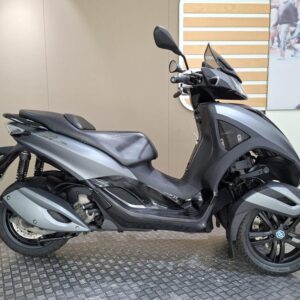 Piaggio MP3 Yourban Sport (2015) A three-wheeled scooter in a sleek silver and black design is displayed on a textured surface against a neutral wall. The scooter features a comfortable seat and modern styling, highlighted by its front and rear wheels.