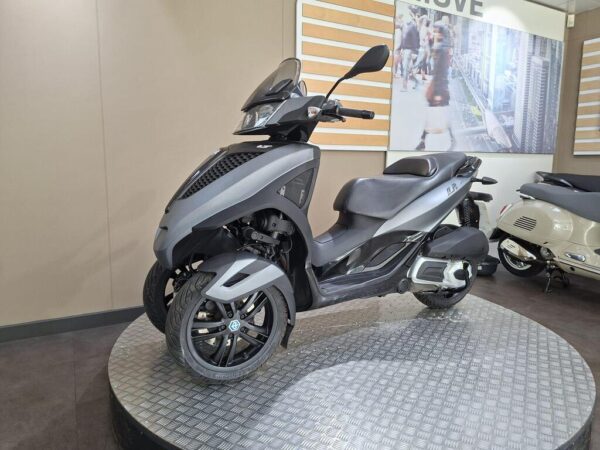 A stylish gray three-wheeled scooter displayed on a circular platform in a modern showroom, with a neutral background featuring a blurred image of urban life and other scooters.