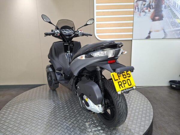 A black three-wheeled scooter displayed on a round metallic platform in a showroom, with a clear view of the front and side. The license plate shows "LF65 RPO."
