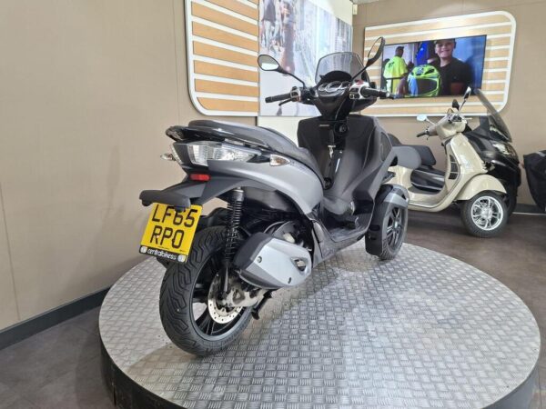 A gray scooter displayed on a circular platform in a showroom, with a license plate reading LF65 RPO, and a blurred background featuring a monitor and another scooter.