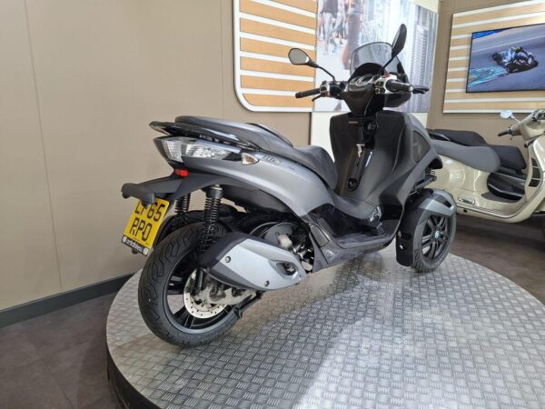A gray scooter displayed in a showroom, featuring two front wheels and a stylish design, with a backdrop of a wall and a screen showing motorcycle-related content.