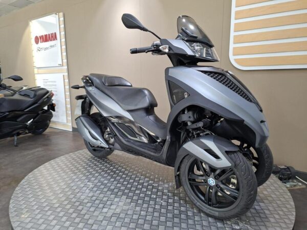 A stylish gray scooter with three wheels displayed on a circular metal platform, set against a neutral background featuring a Yamaha sign.