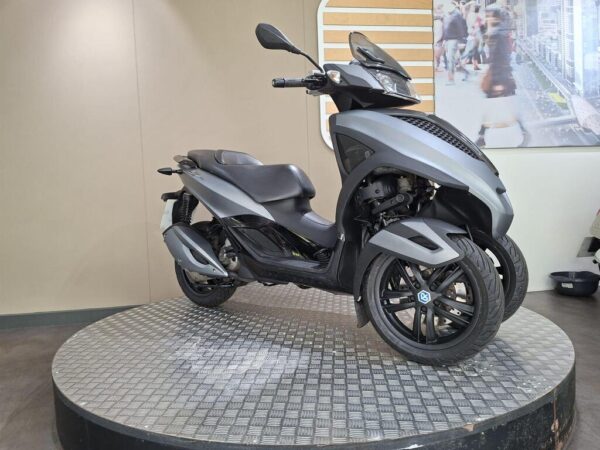 A sleek gray scooter with three wheels displayed on a circular platform in a modern showroom setting. The background features a subtle, light-colored wall and a promotional image of a busy urban scene.