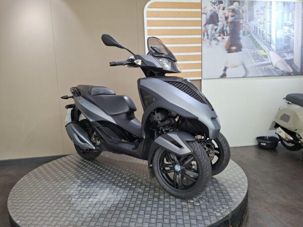 A sleek, gray three-wheeled scooter displayed on a circular platform, set against a neutral background with a promotional image of a city scene on the wall.
