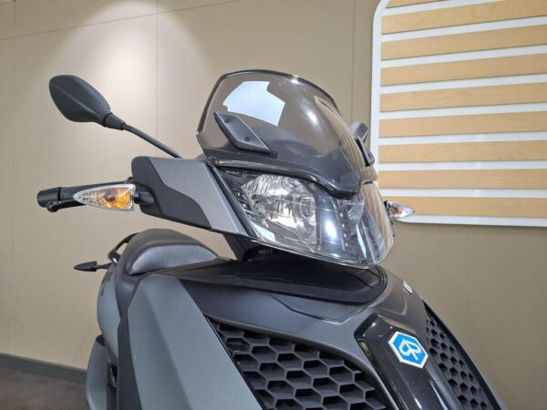 Close-up view of a scooter's front with a clear windscreen, headlight, turn signals, and handlebar reflected on a neutral background.