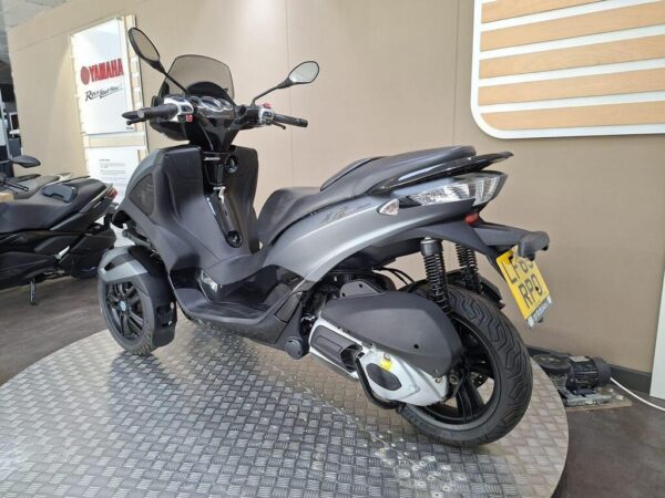 A grey three-wheeled scooter displayed on a textured platform in a Yamaha showroom, with a partial view of another scooter in the background and signage on the wall.
