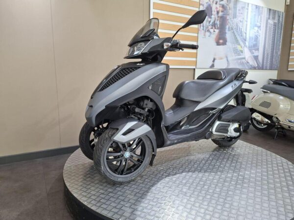 A sleek gray three-wheeled scooter is displayed on a circular platform, featuring a modern design with a front windshield, digital dashboard, and stylish alloy wheels. In the background, a blurred cityscape image is visible, along with another classic scooter.