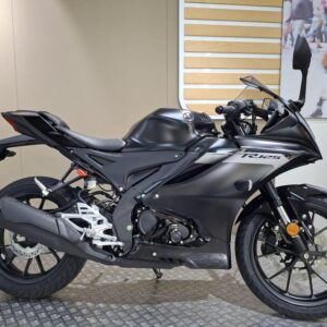 A black motorcycle with a sleek design is displayed on a textured surface, showcasing its sporty features, including a prominent exhaust, modern body shape, and detailed wheels, against a neutral background.