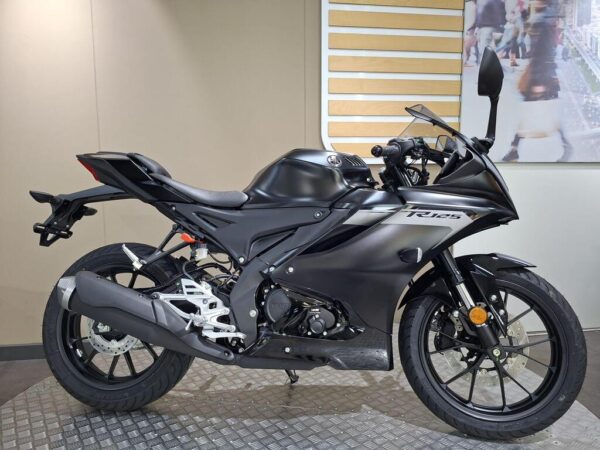 A black motorcycle with a sleek design is displayed on a textured surface, showcasing its sporty features, including a prominent exhaust, modern body shape, and detailed wheels, against a neutral background.