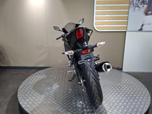 A rear view of a black motorcycle on a circular metallic platform, showcasing its tail lights, exhaust, and tire tread pattern against a neutral background.