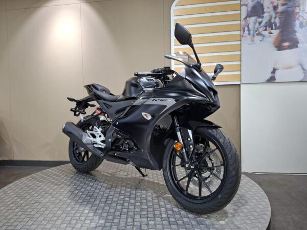 A sleek black sport motorcycle is positioned on a circular platform in a showroom, showcasing its aerodynamic design and modern features. The background features a light-colored wall and blurred figures of people.