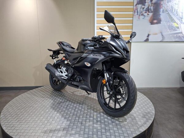 A sleek black motorcycle displayed on a circular metallic platform, with a minimalistic backdrop featuring a blurred image of people walking.