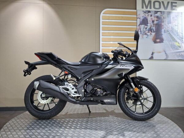 A sleek black motorcycle displayed on a circular metal platform in an exhibition space with a light-colored wall and a promotional background featuring people walking.