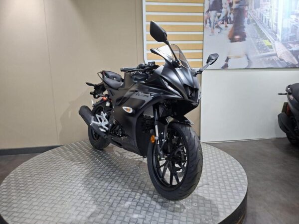 A sleek black motorcycle is displayed on a circular platform, showcasing its aerodynamic design and glossy finish. The background features a minimalistic wall with angled lines and an image of a bustling urban scene.