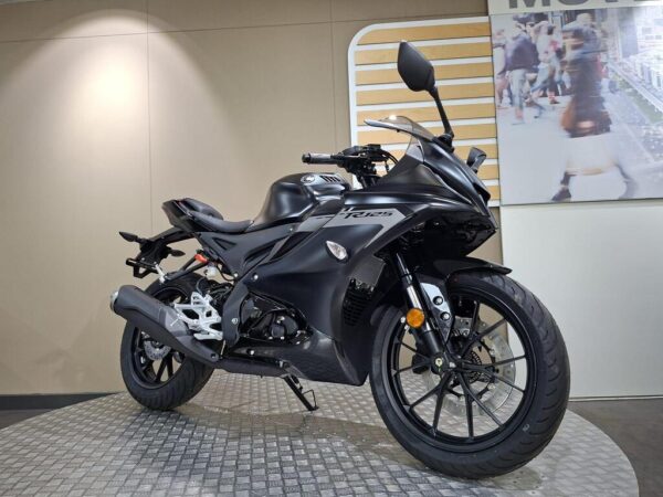 A sleek black motorcycle showcased on a circular platform, featuring a streamlined design with a prominent front fairing, sporty wheels, and modern handlebars. The backdrop includes a subtle wall with a light-colored panel and an image of people in an urban setting.