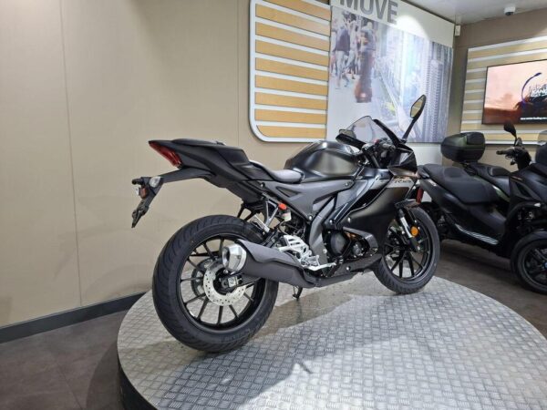 A sleek black motorcycle displayed in a showroom, featuring a modern design and prominent rearview mirrors, with a textured circular platform and a soft backdrop of beige walls and screens.