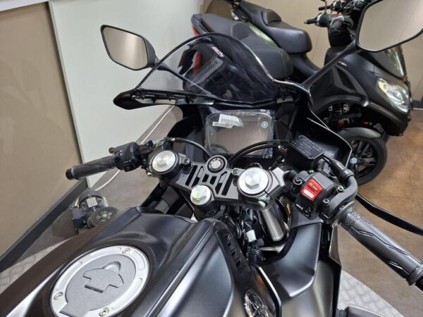 Close-up view of a motorcycle's handlebars, featuring a digital display, control buttons, and mirrors, with additional parked scooters visible in the background.