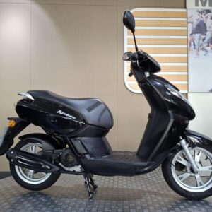 Peugeot Kisbee 50 2018 A sleek black scooter parked on a textured floor, featuring a comfortable seat, modern design, and white wheels, with a stylish background showcasing a blurred urban scene.