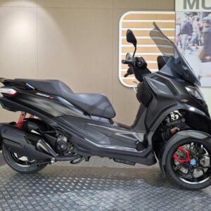 Piaggio MP3 400 (2022) A side view of a sleek, modern black three-wheeled scooter displayed on a circular metal platform, featuring red suspension and accents, set against a minimalistic interior backdrop.