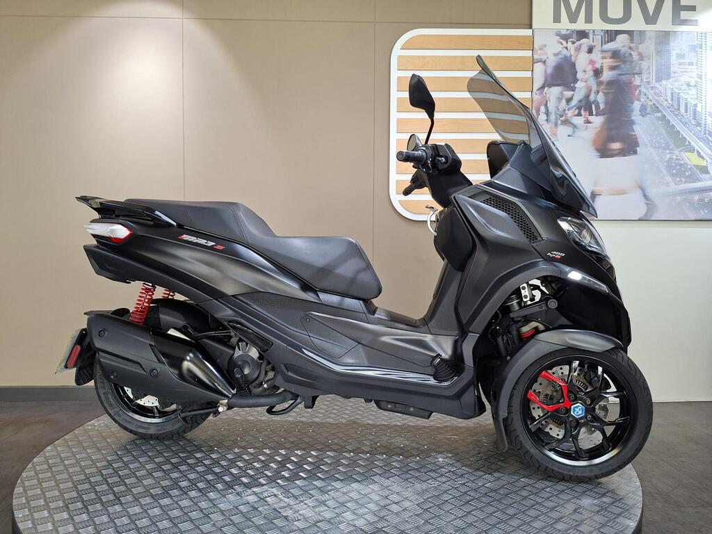 Piaggio MP3 400 LC72 KHB 1 Piaggio MP3 400 (2022) A side view of a sleek, modern black three-wheeled scooter displayed on a circular metal platform, featuring red suspension and accents, set against a minimalistic interior backdrop.