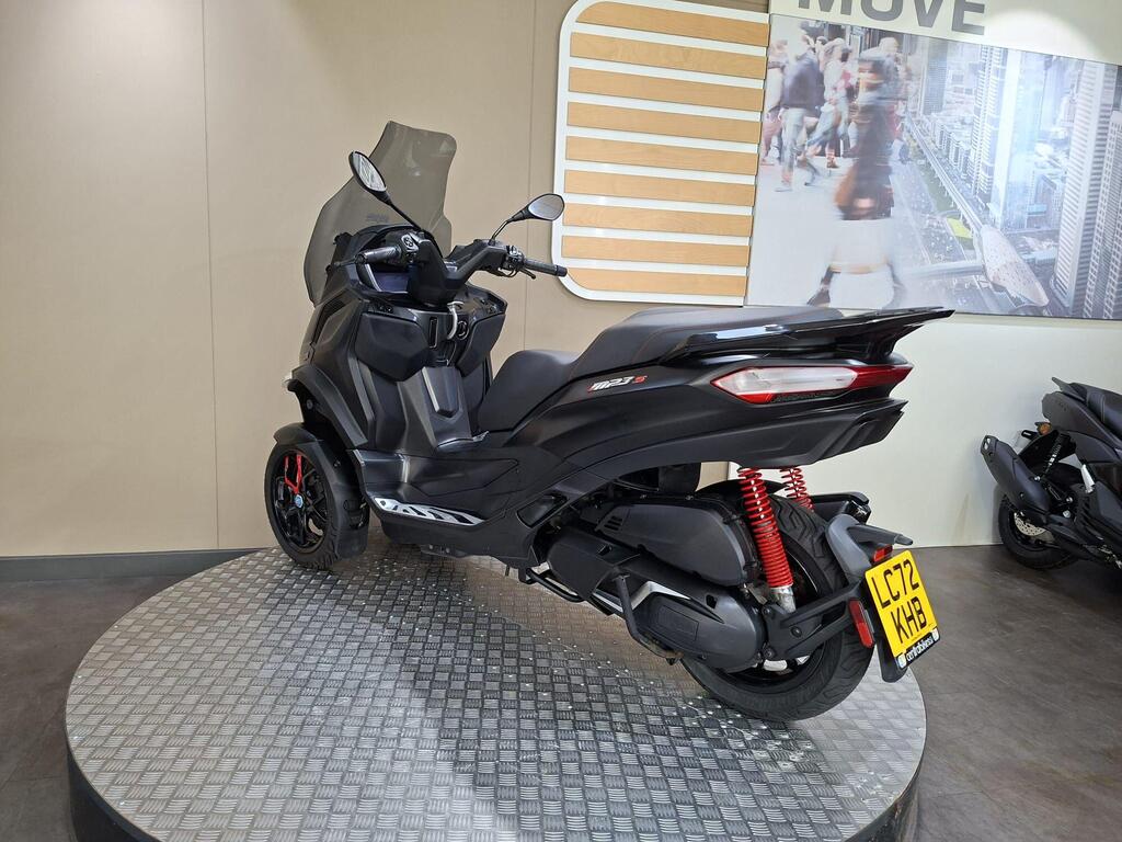 Piaggio MP3 400 LC72 KHB 13 A black three-wheeled scooter is displayed on a circular metallic platform, with a clear windscreen and red shock absorbers, against a light-colored wall and a backdrop featuring a bustling city scene.