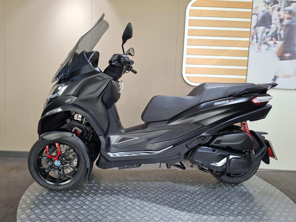 Piaggio MP3 400 LC72 KHB 14 A side view of a black Piaggio MP3 scooter displayed on a patterned gray surface against a neutral background. The scooter features a large windshield, sporty red accents on the wheels, and a sleek design with modern lines.