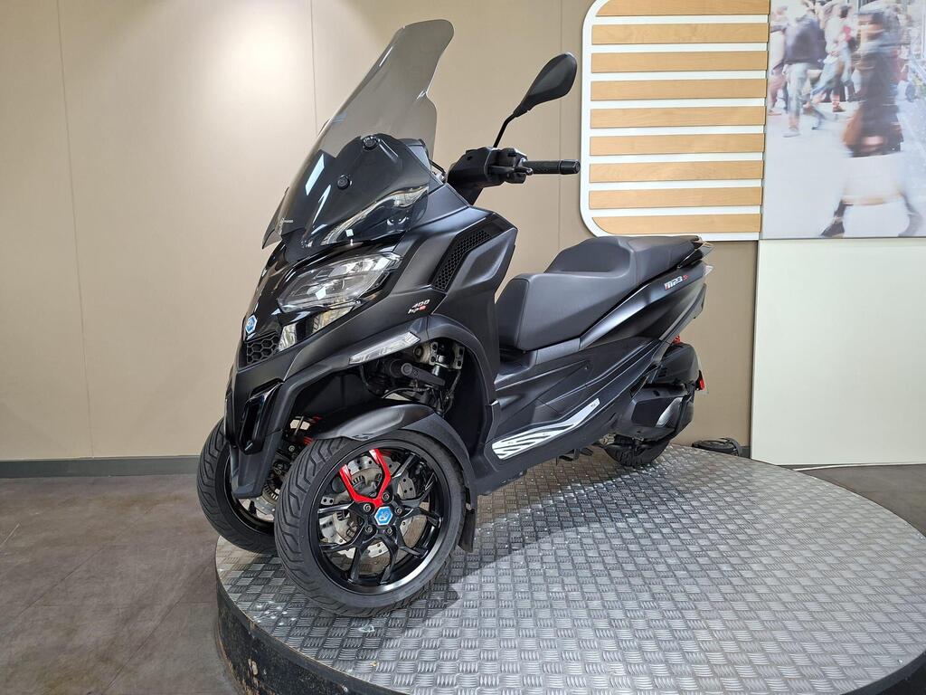 Piaggio MP3 400 LC72 KHB 17 A sleek black three-wheeled scooter with a tinted windscreen, displayed on a circular platform with a textured surface. The scooter features modern design elements, including sporty wheels and a streamlined body. Background includes a large graphic with blurred people.