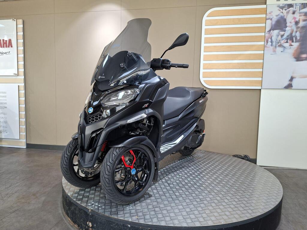 Piaggio MP3 400 LC72 KHB 18 A black three-wheeled scooter with a large windshield is displayed on a round metallic platform in a showroom, with a light-colored wall in the background.